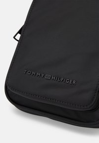 Sort nylon pouch med en glat overflade, featuring en lynlås lukning og præget "TOMMY HILFIGER" logo på forsiden. Slank rektangulær design.