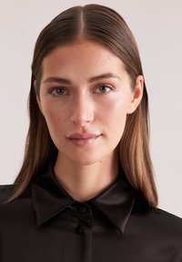 Giovane donna con capelli castani lisci che indossa una camicia nera di satin con bottoni, che guarda direttamente nella telecamera su uno sfondo semplice.