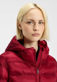 Röd puffjacka med dragkedja framtill och huva. Har en slät, glänsande yta och sydda horisontella paneler.