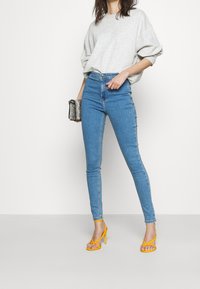 Vaaleanharmaa oversized-huppari, korkea vyötärö siniset skinny-farkut ja keltaiset korot. Asusteina pieni käärmeennahkakuosiset käsilaukku.