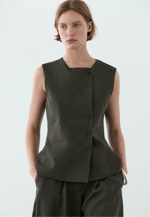 Femme portant un haut sans manches gris foncé boutonné et un pantalon assorti, debout les mains dans les poches devant un fond clair uni.