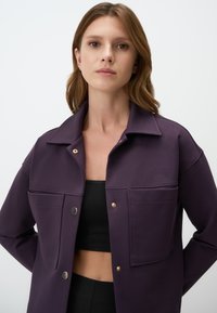 Paarse jas met een puntige kraag, twee voorzakken en goudkleurige drukknoopsluiting, gemaakt van een stevige, geweven stof.