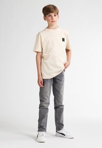 Beige T-shirt met korte mouwen en een zwart logoplakje, gecombineerd met grijze spijkerbroeken en witte sneakers, staande tegen een effen achtergrond.