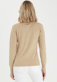 Beige dlouhý svetr s dlouhým rukávem vyrobený z měkkého pleteného materiálu, s těsným střihem a žebrovaným výstřihem. Hladký povrch bez viditelných vzorů.