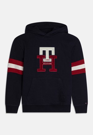 Granatowa bluza z kapturem z przednią kieszenią kangurkową, czerwono-białe paski na rękawach oraz duże czerwono-białe logo "TH Tommy Hilfiger" na piersi.