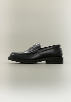 Mocassin penny en cuir noir avec un détail cousu et un petit talon empilé présenté sur un fond clair uni.
