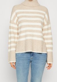 Beżowy i biały sweter z poziomymi paskami, z golfem, noszony z jasnoniebieskimi dżinsami przez stojącą osobę.