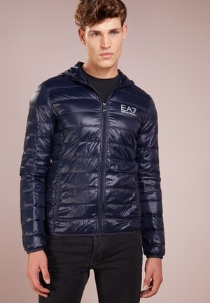 Giacca puffer navy con cappuccio, caratterizzata da un design lucido e trapuntato, zip frontale e logo sul petto. Realizzata in materiale leggero e lucido.