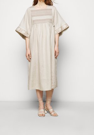 Hellbeiges Leinenkleid mit quadratischem Ausschnitt, weiten Ärmeln und dekorativer Stickerei entlang des Oberteils und der Ärmel. Kombiniert mit Riemchensandalen.