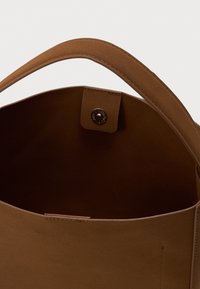 Braune Wildleder-Tote mit einem einzelner Griff, offenem Oberteil und sichtbar innen im Bag einem magnetischen Druckverschluss.