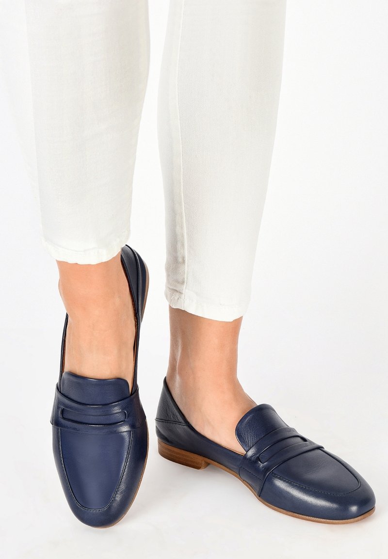 Pieds portant des mocassins en cuir bleu marine assortis à un pantalon blanc coupé au-dessus de la cheville sur un fond blanc.
