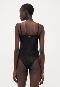 Bodysuit noir en dentelle avec des bretelles fines et ajustables, une texture transparente et un design arrière sans couture. Détail du logo en métal doré dans le haut du dos.