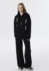 Sudadera negra de gran tamaño con capucha y cordón, presenta múltiples cremalleras metálicas con borlas, combinada con pantalones negros de pierna ancha.