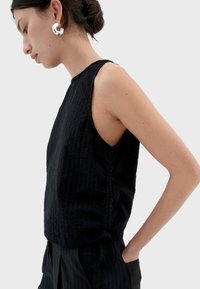 Schwarzes ärmelloses Oberteil mit strukturiertem Finish, featuring einen hohen Halsausschnitt und seitlichen Nahtdetails, kombiniert mit hochtaillierten schwarzen Hosen.