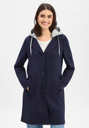 Lächelnde Frau, die einen marineblauen Mantel mit grauem Kapuzenpullover darunter trägt, die Hände in den Manteltaschen vergraben, steht vor einem einfarbigen Hintergrund.