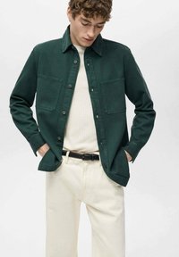 Camicia verde a maniche lunghe con bottoni, due tasche sul petto, colletto strutturato, realizzata in tessuto morbido. Indossata sopra un maglione a coste crema e pantaloni chiari.
