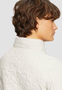 Veste blanche matelassée avec un col haut et des motifs texturés au dos. Tissu lisse avec un design ajusté et élégant.