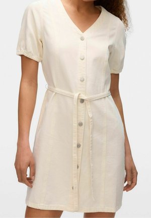 Frau trägt cremefarbenes Kurzarmkleid mit Knöpfen vorne und passendem Stoffgürtel, der in der Taille gebunden ist, und steht vor einem einfarbigen Hintergrund.