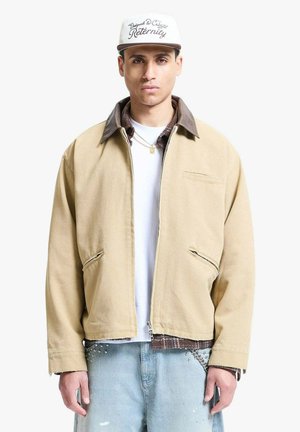 Uomo che indossa una giacca beige con zip sopra magliette a strati, jeans azzurri con borchie, cappellino bianco con testo corsivo e collana d'oro.