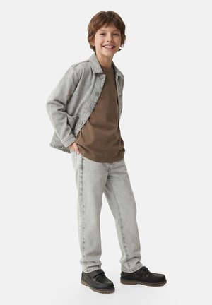 Junger Junge lächelt, trägt eine graue Jeansjacke, braunes Hemd, hellgraue Jeans und schwarze Schuhe mit Riemen, steht vor einem schlichten Hintergrund.