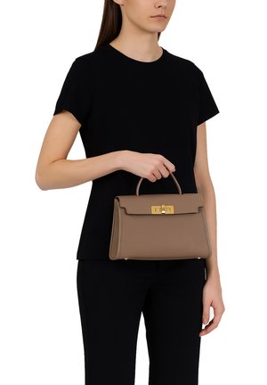 Braune Lederhandtasche mit strukturierter Form, goldfarbener Hardware-Verschluss und kurzem Griff, gehalten von einer Person in schwarzer Kleidung.