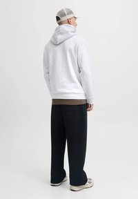 Uomo con felpa grigia chiara, pantaloni larghi neri, sneakers beige e cappellino in rete chiaro in piedi contro uno sfondo semplice, visto da dietro.