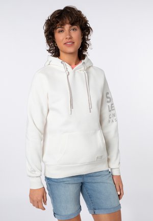 Vrouw met krullend haar die een witte hoodie met voorzak en opgerolde lichtblauwe spijkershorts draagt, staand tegen een effen achtergrond.