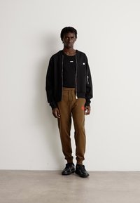 Young man standing against white wall wearing black zip-up jacket, black shirt, brown jogger pants, and black sneakers.