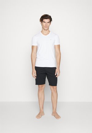 Witte T-shirt met korte mouwen en V-hals, gecombineerd met zwarte shorts. De stof lijkt glad en ademend. Model staat blootsvoets en leunt tegen een effen achtergrond.