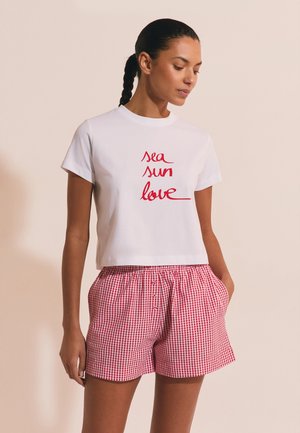 Frau mit geflochtenem Haar, die ein weißes T-Shirt mit rotem "sea sun love"-Schriftzug und rote Vichy-Karo-Shorts trägt, steht mit den Händen in den Taschen.