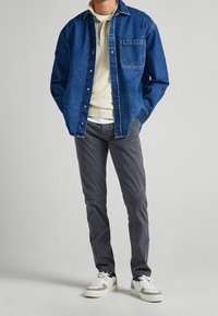 Mężczyzna w niebieskiej jeansowej koszuli z guzikami na beżowej koszulce polo, w szarych spodniach i białych sneakersach, stojący z rękami w kieszeniach na jednolitym tle.