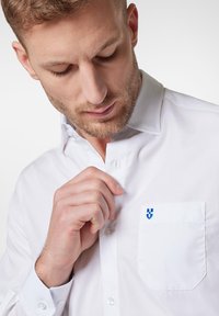 Biała koszula z guzikami, wykonana z teksturowanej tkaniny, z naszywaną kieszonką i małym niebieskim logo. Kołnierz jest usztywniony i wyraźny.