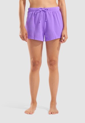 Donna in piedi a piedi nudi che indossa pantaloncini viola in spugna con coulisse su uno sfondo chiaro e semplice.