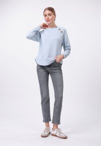 Maglione azzurro chiaro con texture e motivo diagonale, tre bottoni decorativi sulla spalla, abbinato a jeans slim fit grigi e scarpe casual.