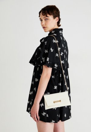 Robe noire avec des motifs de nœuds blancs, manches volantées, associée à un sac à main en cuir blanc rectangulaire avec une bandoulière en chaîne dorée et des ferrures.