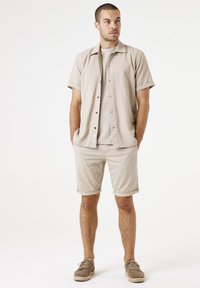 Lichtbeige overhemd met korte mouwen en een gestructureerd patroon, gecombineerd met beige opgerolde shorts en lichtbruine schoenen, wat zorgt voor een gecoördineerde uitstraling.