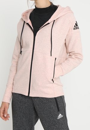 Femme portant un sweat à capuche zippé rose clair Adidas avec des cordons noirs et un pantalon de survêtement Adidas gris foncé.