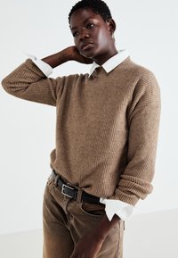 Pull en maille marron en tissu texturé, avec une chemise blanche à col en dessous, porté avec un pantalon marron taille haute.