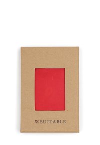 Rood stoffen monster weergegeven in een vierkante uitsparing van een bruine kartonnen verpakking, met het "Suitable" logo in de onderste hoek.