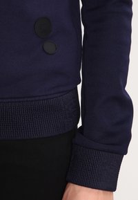 Mörkblå sweatshirt i slätt tyg, med ribbade muddar och fåll, samt två svarta knappar framtill.