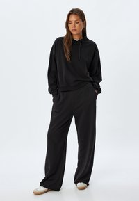 Sudadera negra con capucha, corte relajado, puños y dobladillo acanalados, combinada con pantalones negros de pierna ancha, que cuentan con bolsillos laterales y una textura suave.