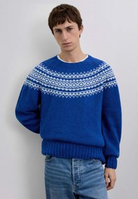 Suéter azul tejido con detalle de canesú estampado en blanco, puños y dobladillo acanalados. Pantalones vaqueros azul claro usados debajo.