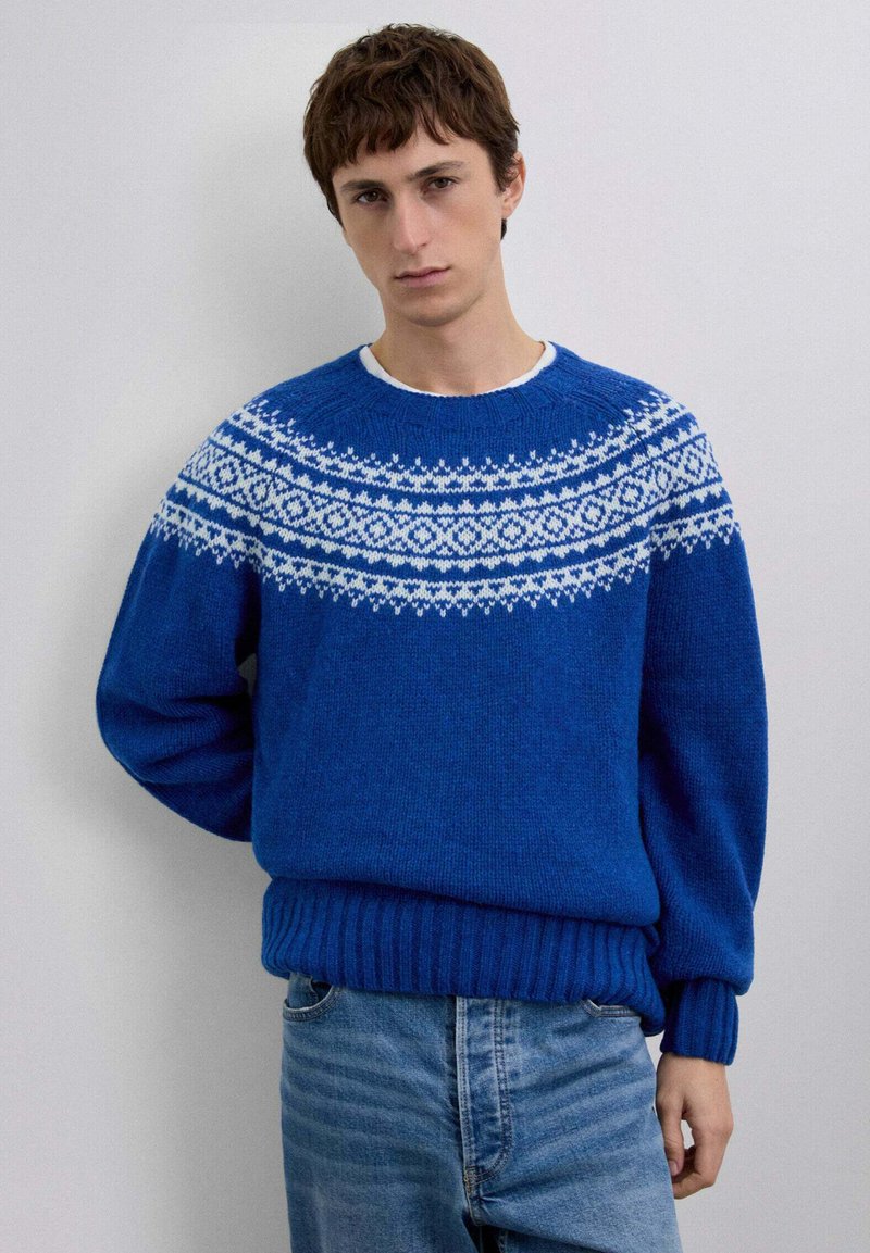 Suéter azul tejido con detalle de canesú estampado en blanco, puños y dobladillo acanalados. Pantalones vaqueros azul claro usados debajo.