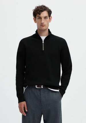 Homme aux cheveux courts et bouclés portant un pull noir à demi-fermeture éclair par-dessus une chemise blanche et un pantalon gris, sur un fond clair uni.