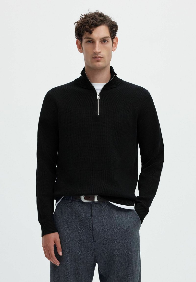 Homme aux cheveux courts et bouclés portant un pull noir à demi-fermeture éclair par-dessus une chemise blanche et un pantalon gris, sur un fond clair uni.