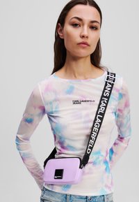 Lilac crossbody bag made of textured material, featuring a black rectangular logo patch and an adjustable strap with bold text.
