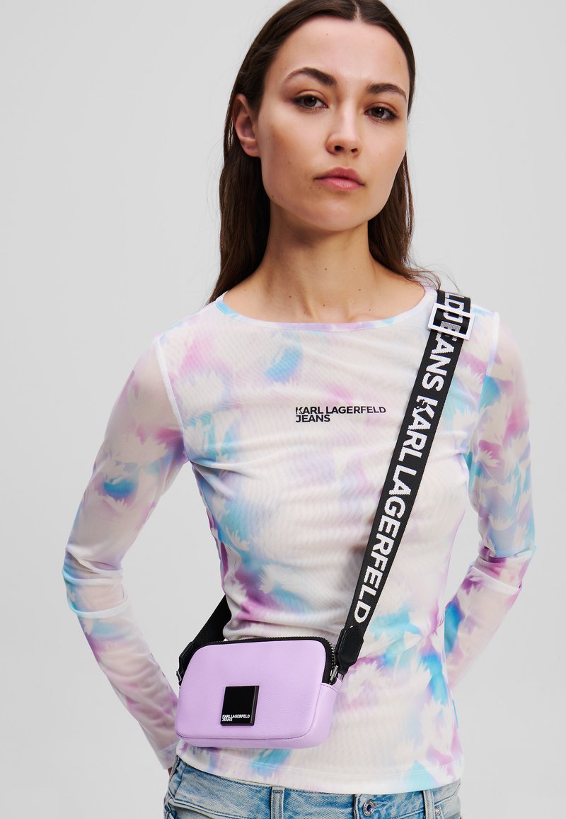 Lilac crossbody bag made of textured material, featuring a black rectangular logo patch and an adjustable strap with bold text.