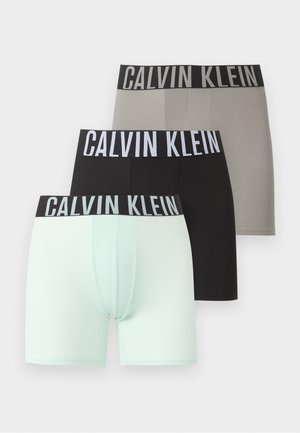 Muskularny czarnoskóry mężczyzna stojący boso, z założonymi rękami, w jasnoszarych bokserkach Calvin Klein na jednolitym białym tle.