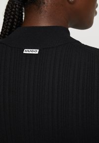 Gros plan sur une personne portant un pull noir côtelé avec une étiquette blanche « HUGO » à l'arrière du col, les cheveux tressés et attachés en arrière.