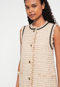 Beige vyšívaná vesta s třepenými okraji, černým lemováním a zlatými knoflíky. Má dvě přední kapsy a kulatý výstřih.